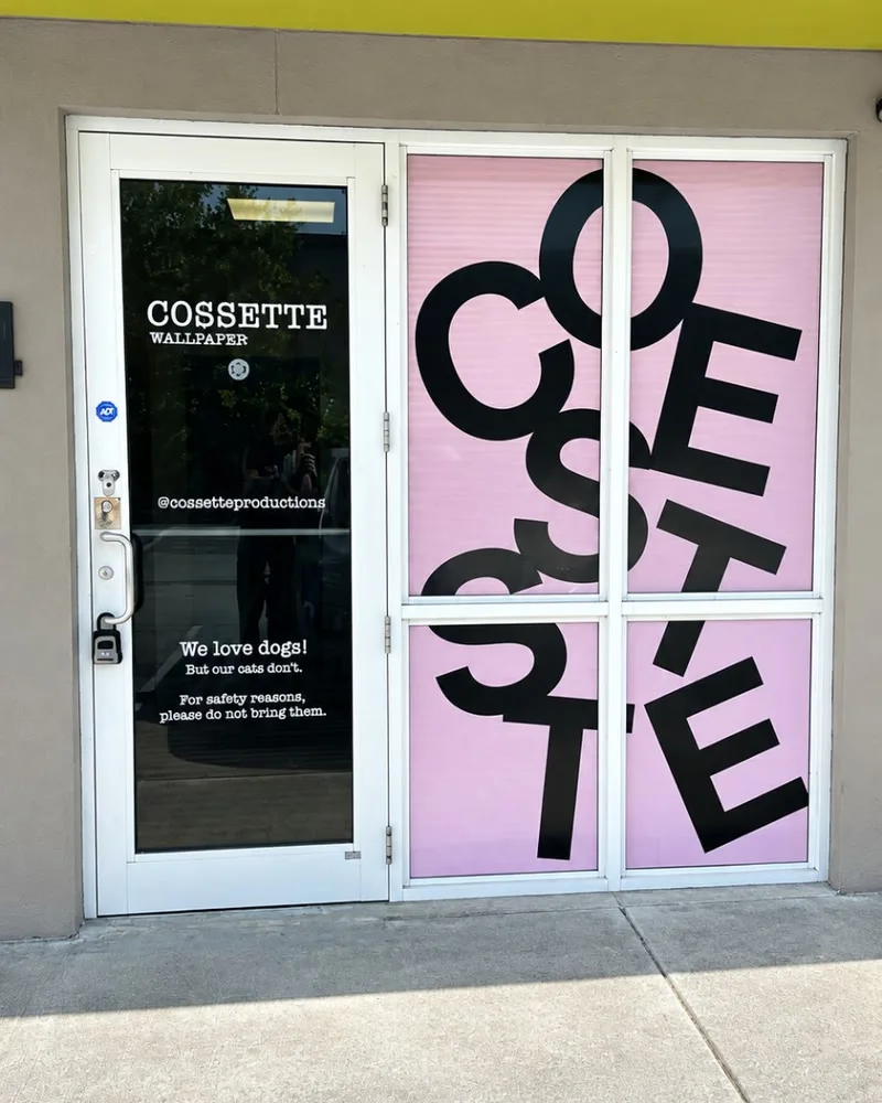 Cossette Wallpaper storefront with giant black COSSETTE letters tumbling across pink window film, beside a glass door with white vinyl lettering, under a lime green awning