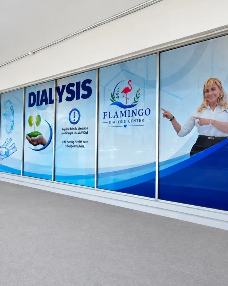 A run of windows at Flamingo Dialysis Center covered in blue wave artwork, with the word Dialysis, the flamingo logo, a message in Spanish and English and a smiling woman pointing at the logo