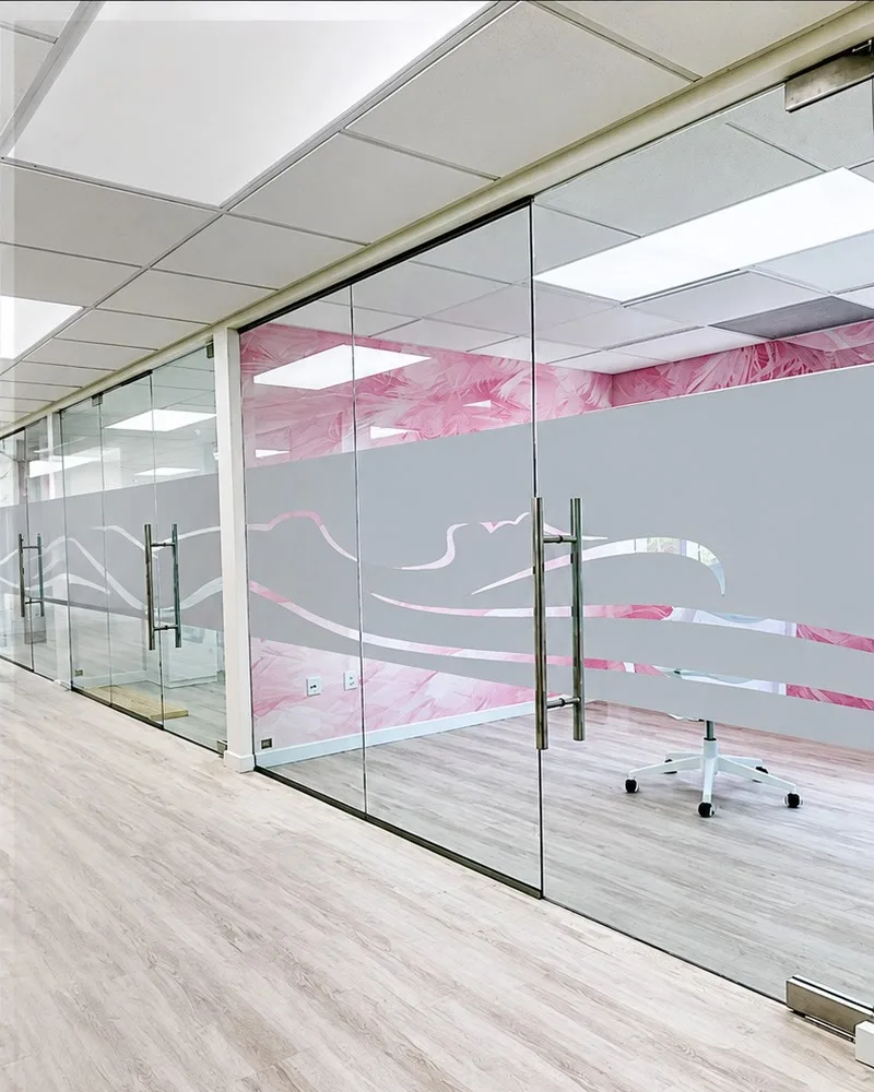 Glass office walls and doors with a frosted vinyl wave band at mid height, above a light wood floor with a pink wall behind the glass